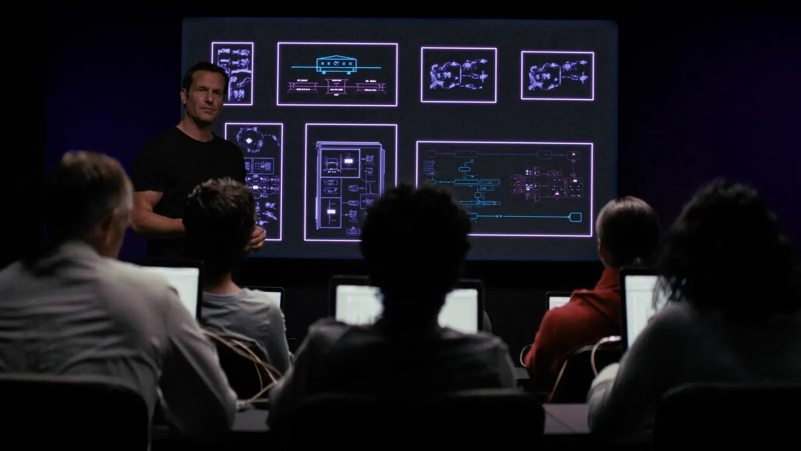 Un instructor explica un diagrama de arquitectura en una pantalla grande ante un grupo con portátiles